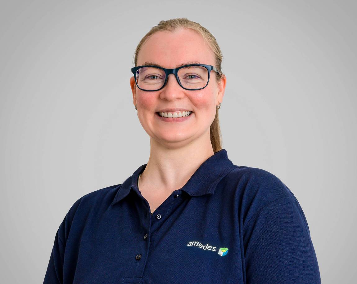 Eine lächelnde Person mit Brille und zurückgebundenen langen Haaren, die ein marineblaues Poloshirt mit dem amedes-Logo trägt, steht vor einem schlichten hellgrauen Hintergrund.
