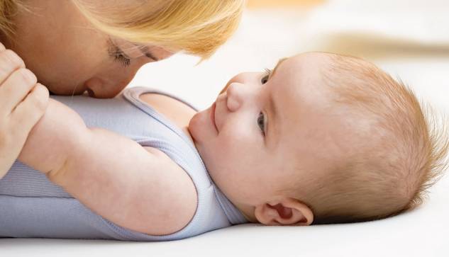 Eine Frau mit blondem Haar küsst sanft die Nase eines lächelnden Babys, das auf dem Rücken liegt, beide auf einer weichen weißen Unterlage. Das Baby schaut die Frau an, die einen hellblauen ärmellosen Strampler trägt.